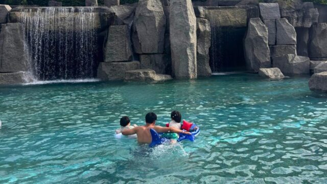 シェラトン・グランデ・トーキョーベイ・ホテルの屋外プールで、岩の滝の前を浮き輪に乗った子ども2人と親が泳ぐ様子