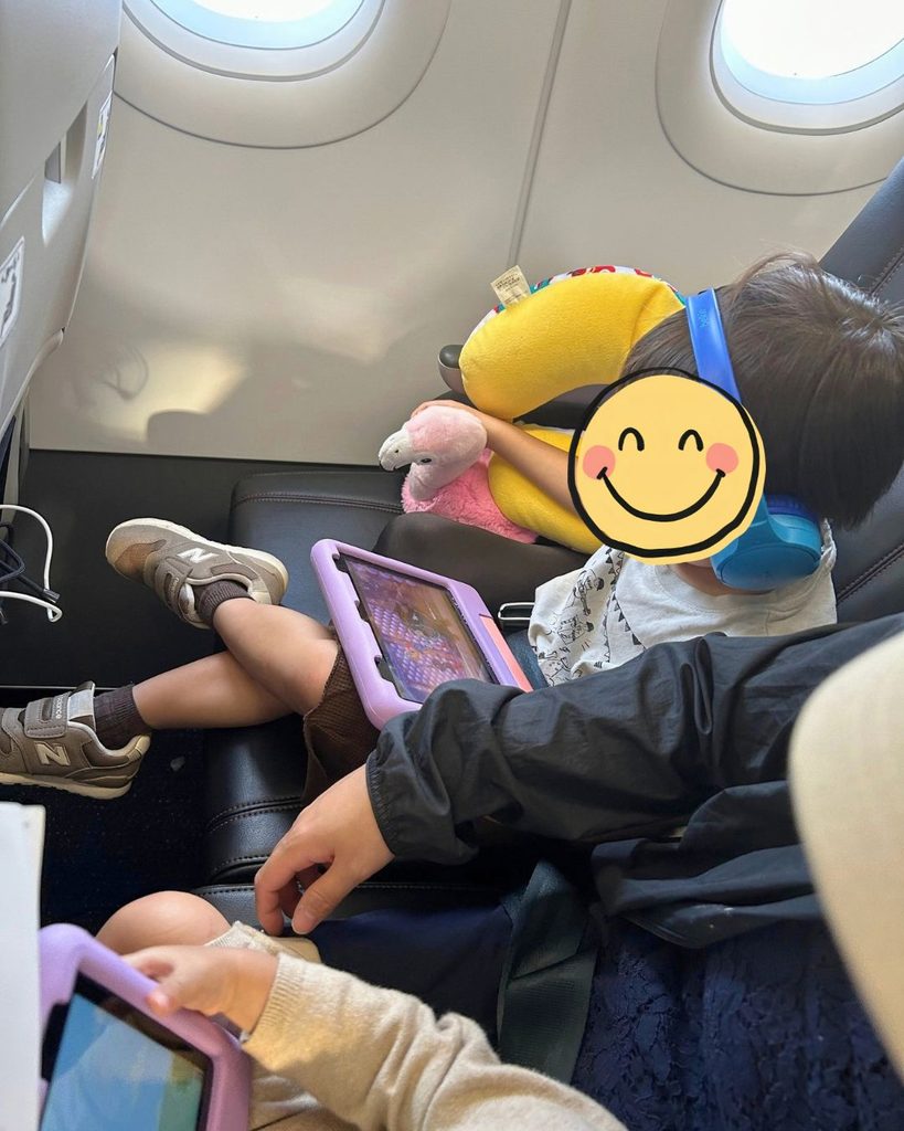 飛行機の座席で、子ども用ヘッドホンをつけてキッズケース付きのタブレットを見る子ども